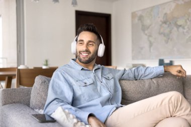 Huzurlu, beyaz kulaklık takan İspanyol bir adam modern koltukta rahatça oturuyor, favori müziğin tadını çıkarıyor, podcast dinliyor ya da sesli kitap dinliyor, evde vakit geçiriyor. Rahatlama, hobi, dinlenme.