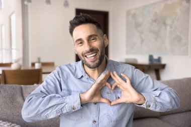 Kanepede oturup kameraya bakan, neşeli, bekar bir adamın portresi birleşmiş parmaklarıyla kalp hareketi yapıyor, bağı, minnettarlığı veya takdiri sembolize ediyor. Aşk ilanı, hayırseverlik.