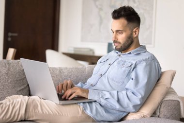 Ciddi Hispanik adam kanepeye yerleşti kablosuz bilgisayar, daktilo, tele çalışma, eşya seçme, otel rezervasyonu, bilet alma, öğrenim, yeni modern platformlar sayesinde yeni bilgiler edindi.