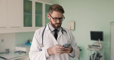 Gözlüklü ve üniformalı genç doktor hastane muayene odasında cep telefonuyla yazı yazıyor, hastayla sohbet ediyor, online danışmanlık yapıyor, tıbbi danışmanlık yapıyor.