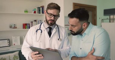 Ciddi bir beyaz doktor, erkek Latin hastaya tıbbi tanı raporunu açıklıyor. Belgeleri kağıt üzerinde gösteriyor, tokalaşıyor, insanlarla tokalaşıyor, mutlu oluyor, gülümsüyor.