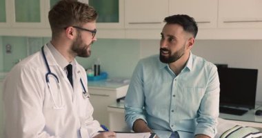 Genç doktor konsültasyonda hastayla konuşuyor, başvuru formu dolduruyor, tıbbi kayıtları tutuyor, adamın şikayetlerini dinliyor, tavsiye veriyor, olumlu düşünüyor, gülüyor, el sıkışıyor.