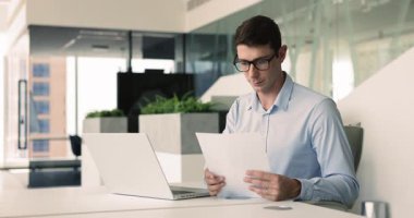 Ciddi genç İspanyol işadamı iş yerindeki kağıt satış raporlarını analiz ediyor, yasal mali belgeleri inceliyor, dizüstü bilgisayarda çalışıyor, muhasebe işi için bilgisayar kullanıyor.