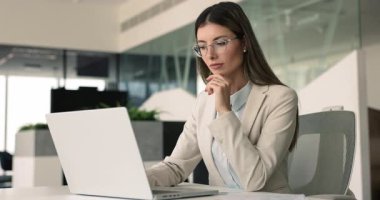 Modern ofiste bilgisayar başında çalışan gözlüklü, düşünceli, ciddi İspanyol şirket CEO 'su kadın. Genç bayan iş adamı dizüstü bilgisayarda yazıyor, çeneye dokunuyor, başka yere bakıyor, yaratıcı projeyi düşünüyor.