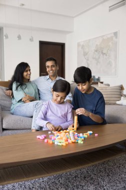 Genç Hintli ebeveynler kanepede oturur, küçük oğulları ve kızları masada oturup plastik yapı bloğu oynarken, aile hafta sonunu evde geçirir, boş zamanlarının ve oyun zamanlarının keyfini çıkarırlar.