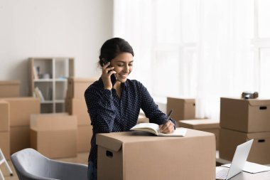 Sipariş alıyorum. Gülümseyen genç iş kadını girişimci serbest pazarlamacı web market sahibi stoklardaki ofis masasından müşteriye telefonla konuşuyor anlaşma notlarını kağıt deftere not alın.