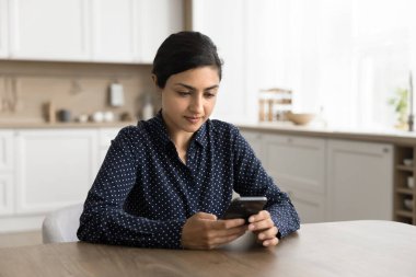 Çevrimiçi iletişim. Milenyum kadın serbest meslek sahibi. Mutfak masasında oturup akıllı telefon ekranına odaklanmış. Müşteriye mesaj yazıp sosyal ağ sayfalarını okusunlar.