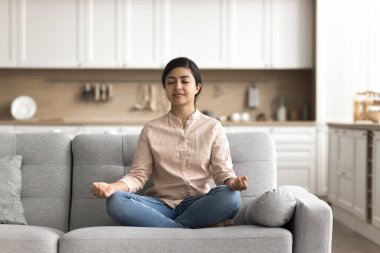 Meditasyon zamanı. Sakin, sakin genç bir kadın modern evdeki rahat kanepeye oturur yoga üzerine yoğunlaşır, gözleri kapalı meditasyon yapar jimnastik yapar, uyumunu hisseder duygusal dengeyi rahatlatır.
