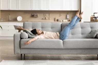 Boş zaman. Heyecanlı, kaygısız genç kadın rahat bir kanepeye uzanıyor ve arka planda modern iç mimari ile birlikte hafta sonları evde özgürlüğün tadını çıkarıyor işten sonra yorgun bacaklara istirahat veriyor ev işleri bitince dinleniyor.