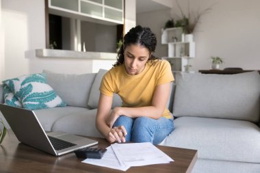 Ciddi Latin kadın hesap makinesi kullanarak kanepeye oturur, faturaları laptopta e-banka kullanarak öder, kişisel bütçeyi yönetir, gelir ve kontrol eder, muhasebe işlerini evde yapar.
