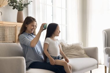 Sağlıklı sabah rutini. Sevgi dolu genç anne dadı, oturma odasındaki koltukta oturmadan önce uzun saçlı, kreş kızlarını silikon fırçayla fırçalayan küçük kızla ilgileniyor. Boşluğu kopyala