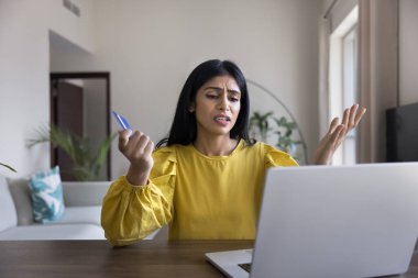 Hintli kadın elinde kartla dizüstü bilgisayar ekranına bakıyor, ödemeyi başarısız yapmaya çalışıyor, çıkış sırasında ek ücret veya nakliye masrafları nedeniyle endişeli hissediyor, kart limiti