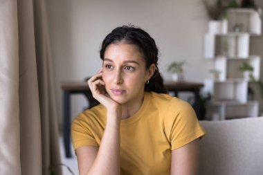 Sarı günlük tişört giymiş güzel bir kadın kanepeye oturmuş, çenesini açmış, hafif bir gülümsemeyle mesafeye bakıyor, evde tek başına vakit geçirmenin tadını çıkarıyor.