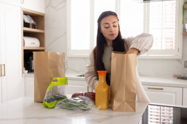 Güzel, genç bir ev hanımı mutfak masasında kağıt süpermarket poşetlerinden yiyecek üretiyor, organik yiyecek ve taze sebze paketlerini açıyor.