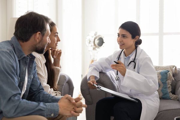 Позитивная молодая индийская женщина-врач, посещающая пару дома, записывающая медицинские записи, заполняющая информацию о пациентах, разговаривающая с мужем и женой, улыбающаяся, дающая оптимистичные консультации