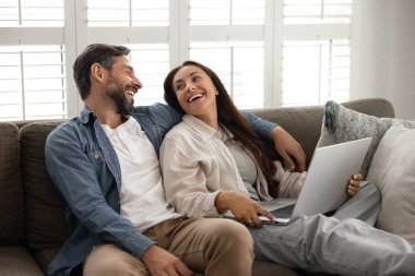 Mutlu, neşeli Latin çift evlerinde internet iletişiminden keyif alıyor, online TV multimedya servisini kullanıyor, evinde rahat bir kanepeye oturmuş, aletlerle konuşuyor, gülüyor, gülüyor.