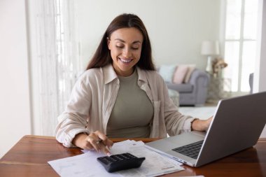 Evde hesap makinesine yazı yazan, bütçeyi hesaplayan, geliri, birikimleri, faturaları ödeyen, dizüstü bilgisayardaki online finansal uygulamadan vergi alan, finansal yönetimden keyif alan genç, pozitif ve serbest çalışan bir kadın.