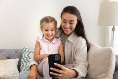 Neşeli genç bir anne kucağında tatlı küçük bir kızı tutuyor, akıllı telefonuyla selfie çekiyor, cep telefonunu internet iletişimi için kullanıyor, video konuşmaları yapıyor, boş zamanların tadını çıkarıyor, aile eğlencesi yapıyor.