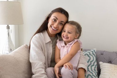 Mutlu, genç bir anne kucağında tatlı bir kız çocuğu, rahat bir kanepede rahatlıyor, aile eğlencesinin, anneliğin tadını çıkarıyor, kameraya bakıyor, gülümsüyor, gülüyor, resim için poz veriyor.