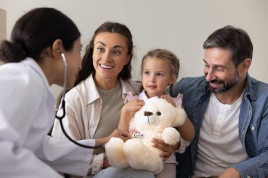 Genç Hintli doktor, evde kucağında oturan küçük kızı muayene ediyor. Çocuk doktoru hasta çocukla temas ve güven için oynuyor, oyuncak ayıların göğsünü dinliyor.