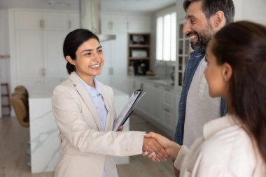 Neşeli genç Hintli emlakçı kadın mutlu ev sahipleriyle tokalaşıyor, belgeleri tutuyor, gülümsüyor. Başarılı daire avcılığı için teşekkür eden çift emlakçı.