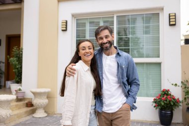 Neşeli orta yaşlı bir çift emlak alıcısı yeni evin önünde poz veriyor, ev sahipliğini kutluyor, dışarıda dikiliyor ve sarılıyorlar.