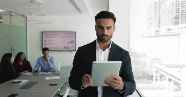 Resmi takım elbiseli işadamı konferans odasında dijital tablet kullanarak duruyor, çevrimiçi raporları inceliyor, alınan verileri analiz ediyor veya görevleri yönetiyor. Yenilikçi teknoloji, modern aygıt kullanarak iş akışı