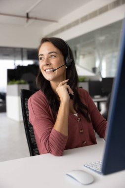 Kulaklık takan, bilgisayar monitöründeki müşteriyle konuşan, yüzünü çeviren, gülümseyen, müşteriyle çevrimiçi iletişim kurmayı düşünen bir kadın.
