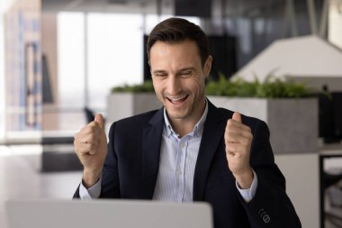 Heyecanlı yakışıklı Latin iş adamı zaferden, zaferden, laptopta çalışmaktan zevk alıyor, el kazanmasını sağlıyor bilgisayarlara el hareketi yapıyor, gülüyor, ekrana bakıyor, başarıyı kutluyor.