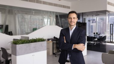 Mutlu yakışıklı İspanyol iş adamı modern şık ofis çalışma alanında tek başına duruyor, kollarını kavuşturmuş kendine güvenen hareketlerle poz veriyor, kameraya bakıyor, gülümsüyor. Ön afiş portresi