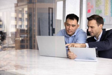 İki farklı iş adamı online proje yönetimini tartışıyor, tartışma için dizüstü bilgisayar ve tablet kullanıyorlar, ekrana işaret ediyorlar, konuşuyorlar, birlikte internet girişimi üzerinde çalışıyorlar.