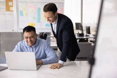 Mutlu beyaz erkek patron, Asyalı yöneticiyi kontrol ediyor laptopta yazı yazıyor, tavsiye veriyor. Neşeli iş arkadaşları oturur, oturur, konuşur, bilgisayara güler.