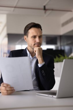 Belgelerle çalışan odaklanmış Latin işadamı, iş yerindeki raporları kontrol ediyor, kağıt mektubu tutuyor, dizüstü bilgisayar kullanıyor, çeneye dokunuyor, düşünüyor, karar veriyor, bilgisayardaki metni okuyor.