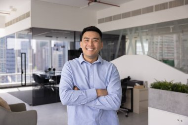 Kurumsal ofis alanında tek başına kurumsal profesyonel portre için poz veren kendine güvenen Asyalı bir yönetici. El ele tutuşup güven jesti yapıyor ve dişlek bir gülümsemeyle kameraya bakıyor.