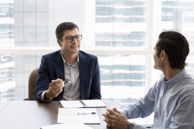 Beyaz iş adamı resmi takım elbiseli, şirket müşterisiyle görüşmelere öncülük ediyor, masada birlikte oturuyor, gelecekteki işbirliğini planlıyor, şirket hizmetlerini satıyor, ikna ediyor, açıklamalar yapıyor.