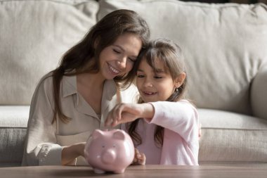 Gelecek için biriktiriyorum. Mutlu gülümseyen bin yıllık anne küçük kız öğrenci oturma odasında sarılıp kumbaraya bozuk para atıyor. Finansal bilgi bankası rasyonel ekonomi kavramı