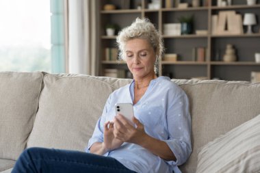 Zamana ayak uyduruyorum. Güzel stil sahibi yaşlı kadın emekliler modern oturma odasındaki kanepeye oturur akıllı telefon hesabındaki web uygulamasını kullanır online banka hesabındaki mesajları sosyal medya üzerinden okurlar.
