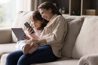Dijital teknoloji ve aile zamanı. Mutlu bekar anne kız çocuğu kanepeye kıvrılıp çeşitli hesaplardan internet oyunu oynayarak veri paylaşımı yapan aletlerle eğleniyor. Boşluğu kopyala
