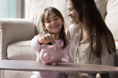 Finansal alışkanlıklar edinmek. Gülümseyen genç bir anne, küçük mutlu kızının domuzcuk kumbarasına para koymasına destek oluyor. Aile finans eğitimi yurt içi ekonomi para yönetimi kavramı. Boşluğu kopyala