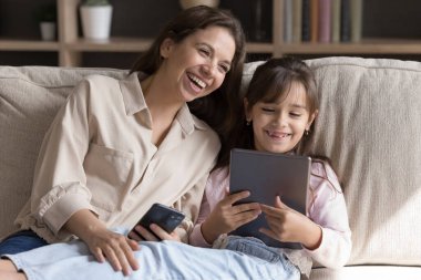 Samimi kahkaha ve dijital bağlantı. Neşeli yetişkin abla ve küçük kız kardeş birlikte gülüyorlar kanepede oturuyorlar ve sosyal medyada komik fotoğrafları karşılaştırıyorlar.