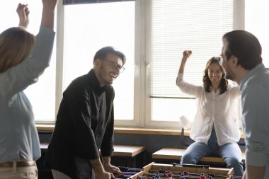 Neşeli milenyum işadamı masa futbolunda iş arkadaşını dövüyor. Mutlu iş arkadaşları iş molasında eğleniyor, oyuncak futbolda yarışıyor, gülümsüyor, gülüyor, iki oyuncuya destek oluyor.