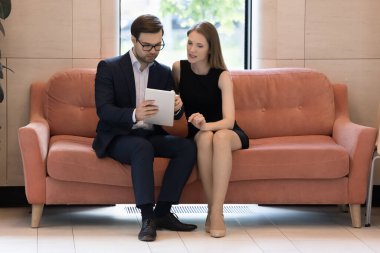 Çalışma odasındaki koltukta çalışan iş arkadaşları dijital tablet kullanıyorlar. Şirket temsilcisi, sunum yöneticisi, modern cihazda müşteriye ticari teklifte bulunuyor. Modern teknolojiyi kullanarak takım çalışması