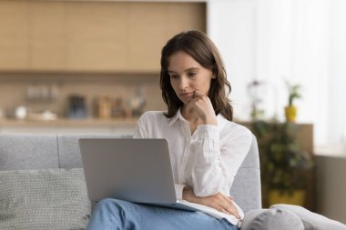 İş iletişimi için online uygulama kullanan, bilgisayar başında çalışan, evdeki koltukta oturan, e-posta mesajlarını okuyan, makale yazan, çeneye dokunan, düşünen düşünceli, düşünceli bir serbest çalışan kız.