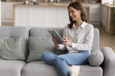 Neşeli, güzel blogger kızı evdeki elektronik cihazda online uygulama kullanıyor, video görüşmesinde konuşuyor, kablosuz iletişimin tadını çıkarıyor, yumuşak kanepeye uzanıyor, tablet tutuyor.