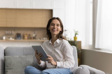 Mutlu güzel tablet kullanıcısı kadın ev portresi. Genç ve güzel bir blog yazarı, elinde dijital bir cihazla koltukta oturmuş, dişlek gülümseyerek kameraya bakıyor, çevrimiçi iletişimin tadını çıkarıyor.