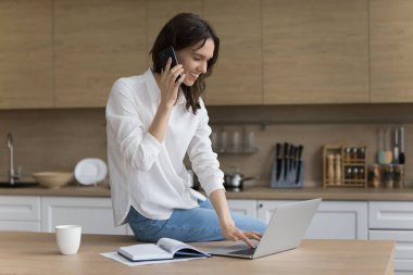Ev işyerinde cep telefonuyla konuşan başarılı serbest çalışan bir kadın, dizüstü bilgisayar ve defterde mutfak masasında oturuyor, iş görevlerini tartışıyor, gülümsüyor, gülüyor, iş iletişiminden keyif alıyor.