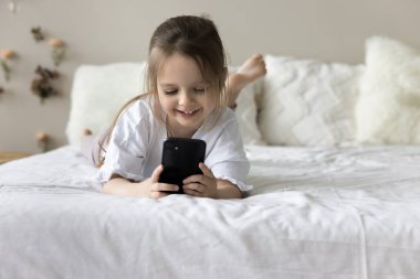 Mutlu tapılası küçük kız cep telefonuyla online öğrenme oyunu oynuyor, beyaz çarşaflı çift kişilik yatakta dinleniyor, akıllı telefonda çizgi film izliyor, internet teknolojisini cihaz üzerinde kullanıyor.