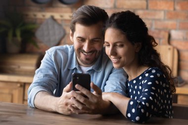 Gülümseyen genç bir çiftin kucaklaşması, akıllı telefon kullanması, mutfaktaki ahşap masada oturması, boş zamanların keyfini çıkarması, alışveriş yapması ya da sosyal ağlarda sohbet etmesi.