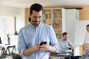 Ofisteki genç beyaz erkek çalışan akıllı telefon ekranındaki kablosuz internete bakıyor. Milenyum çalışanı cep telefonu mesajını cihazda kullanıyor. Teknoloji, iletişim konsepti.