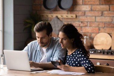 Ciddi genç bir çift mutfakta ev faturalarını hesaplıyor, bütçeyi yönetiyor, dizüstü bilgisayar kullanan kadın ve erkek odaklanıyor, online bankacılık hizmetlerine göz atıyor, vergi ödüyor, mali belgeleri kontrol ediyor.